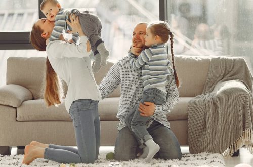 Cheerful mother kissing little boy while father hugging adorable daughter when family sitting on floor in rainy day against window