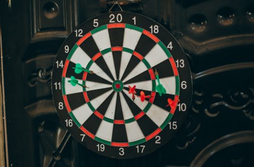 Close-up of a dartboard with red and green darts hitting various spots, indoors setting.